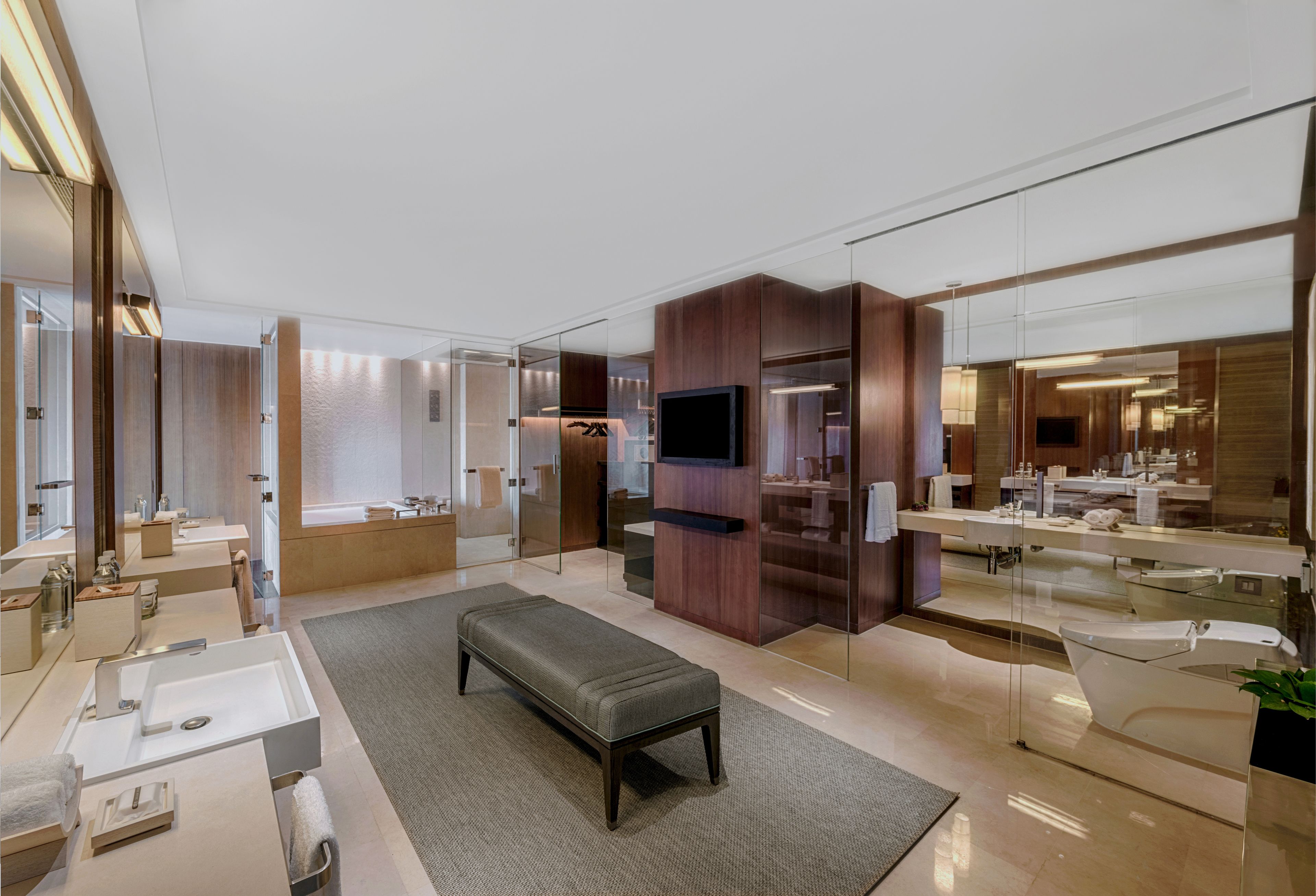 A modern bathroom with a large mirror, multiple sinks, a bench, and a television.