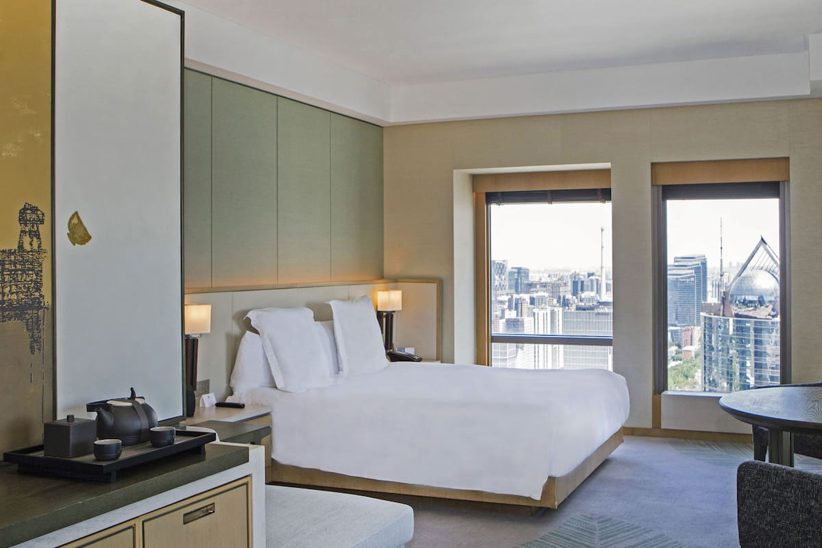 Une chambre d’hôtel avec un lit, une table de chevet, une lampe et une vue sur le paysage urbain grâce à de grandes fenêtres.