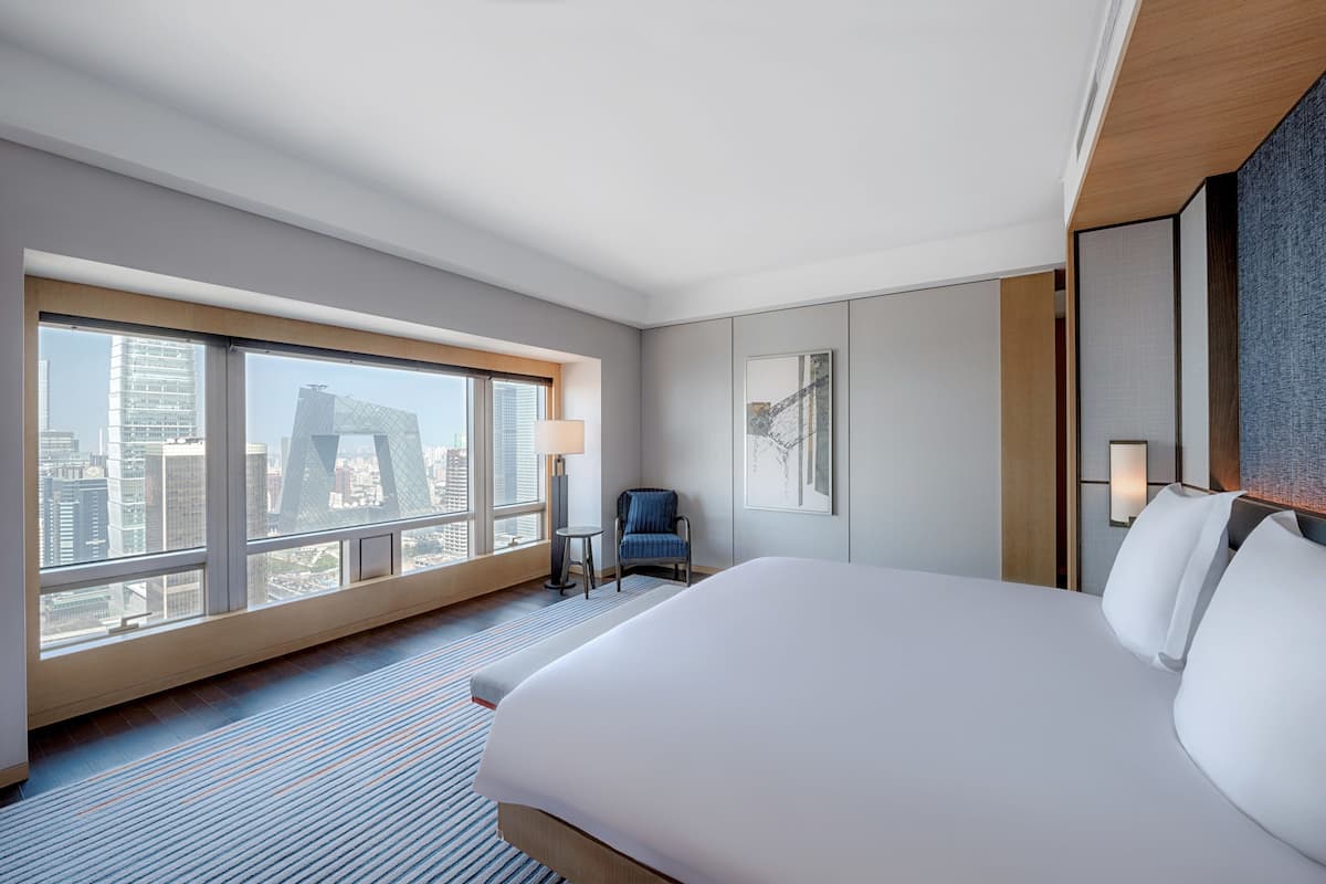 Une chambre d’hôtel avec un grand lit, une chaise, une petite table et une vue sur la ville grâce à de grandes fenêtres.