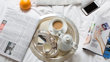 A tray with a teapot, cups, and a glass of water on a bed with newspapers and a magazine.