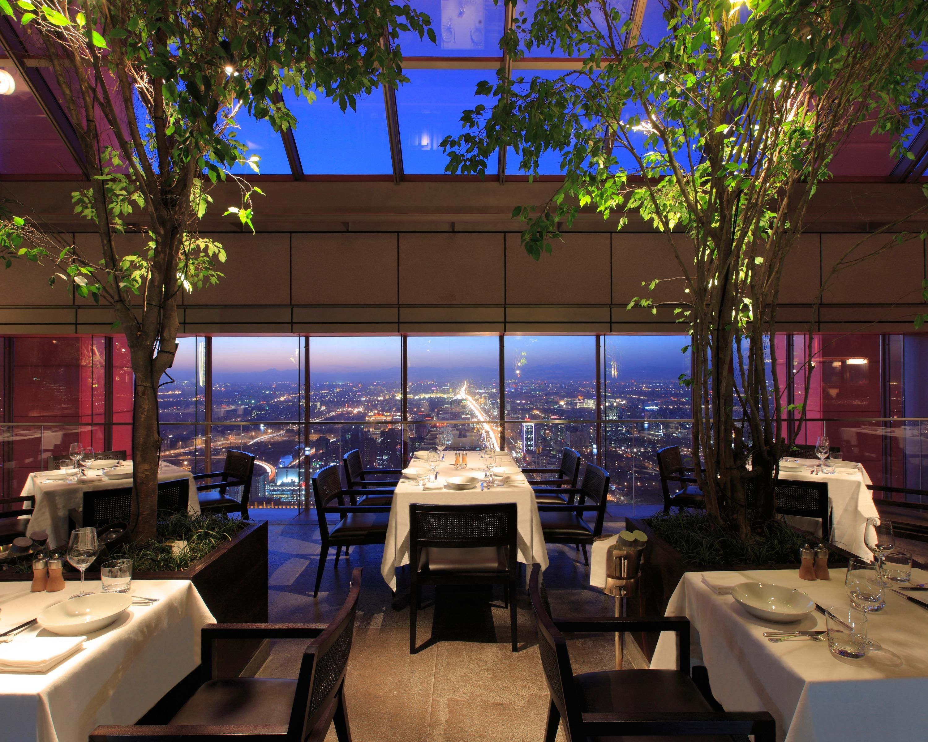 An indoor dining area with tables set for two, large windows offering a cityscape view, and trees providing natural light.