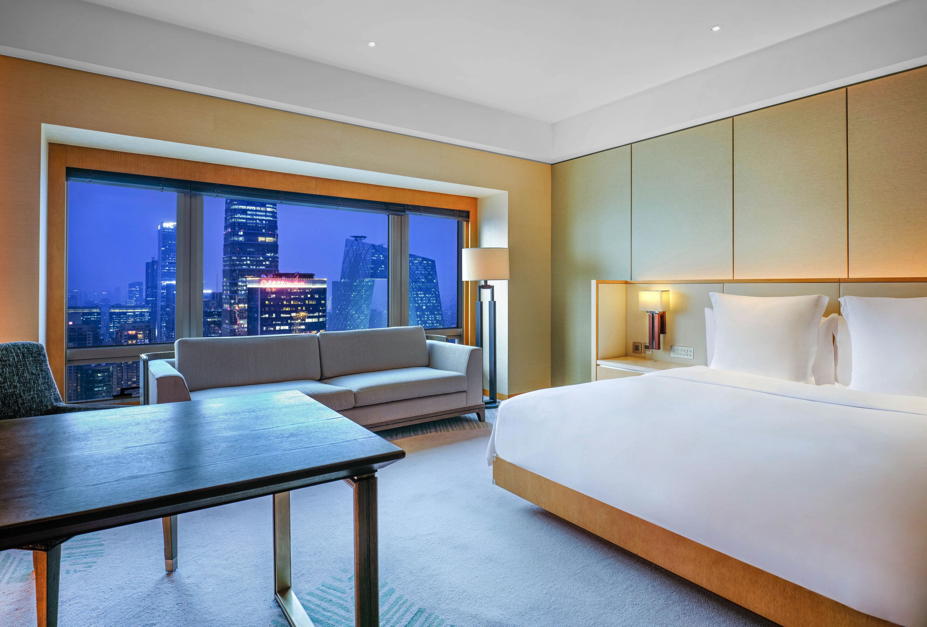 Camera d'albergo moderna con un grande letto, un divano, un tavolo da pranzo e vista sullo skyline cittadino di notte.