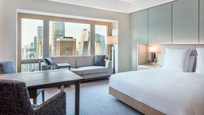 Habitación de hotel con una cama grande, un sofá, una mesita y una silla. Cuenta con un ventanal con vistas a la ciudad.