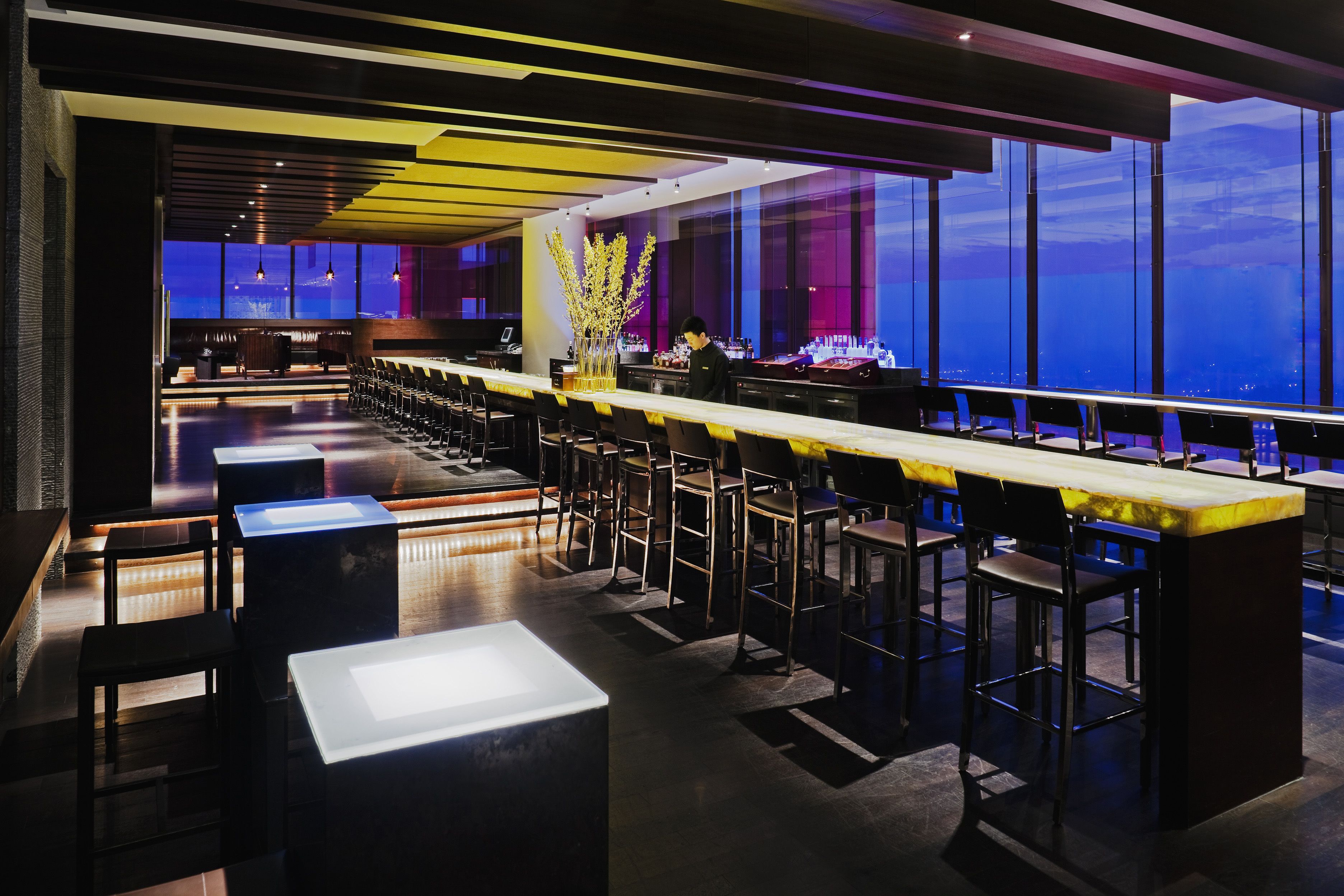A modern bar area with high tables and stools, a large vase with branches, and a view of the city at night.