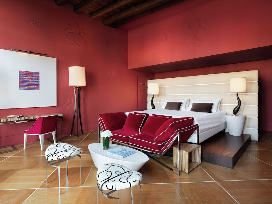 A modern hotel room with a red accent wall, a bed with white bedding, a red sofa, and a white coffee table.