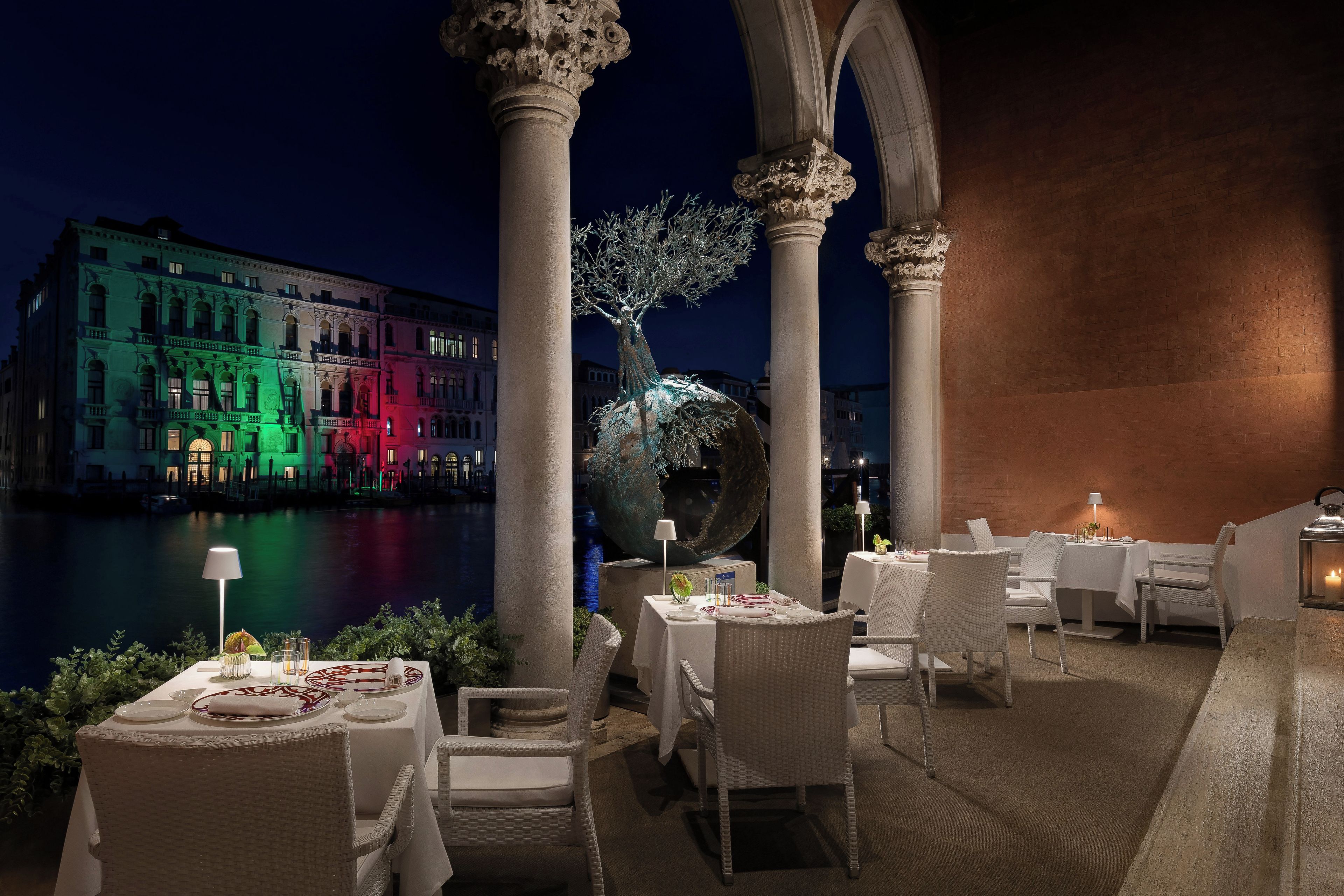 Un espace de restauration en plein air avec des tables et des chaises, une sculpture d’arbre décorative et une vue sur un bâtiment dont les fenêtres sont illuminées.