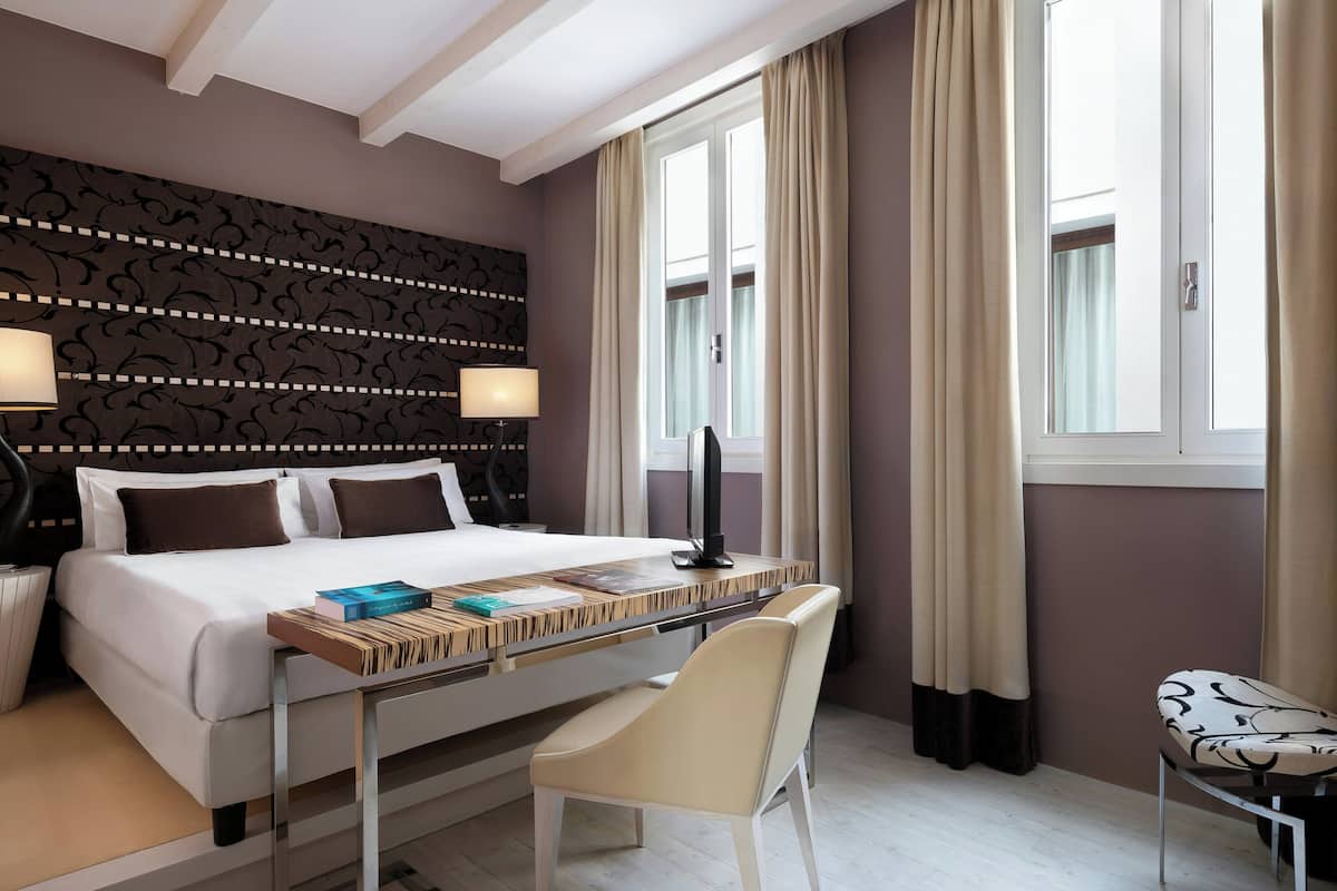 Una habitación de hotel con cama, mesita de noche, lámpara, silla y escritorio con libros.