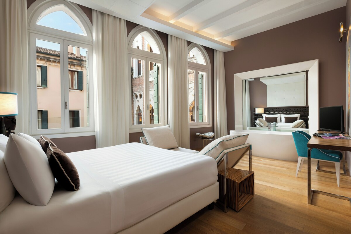 Habitación de hotel con cama, escritorio, silla y ventanas amplias.