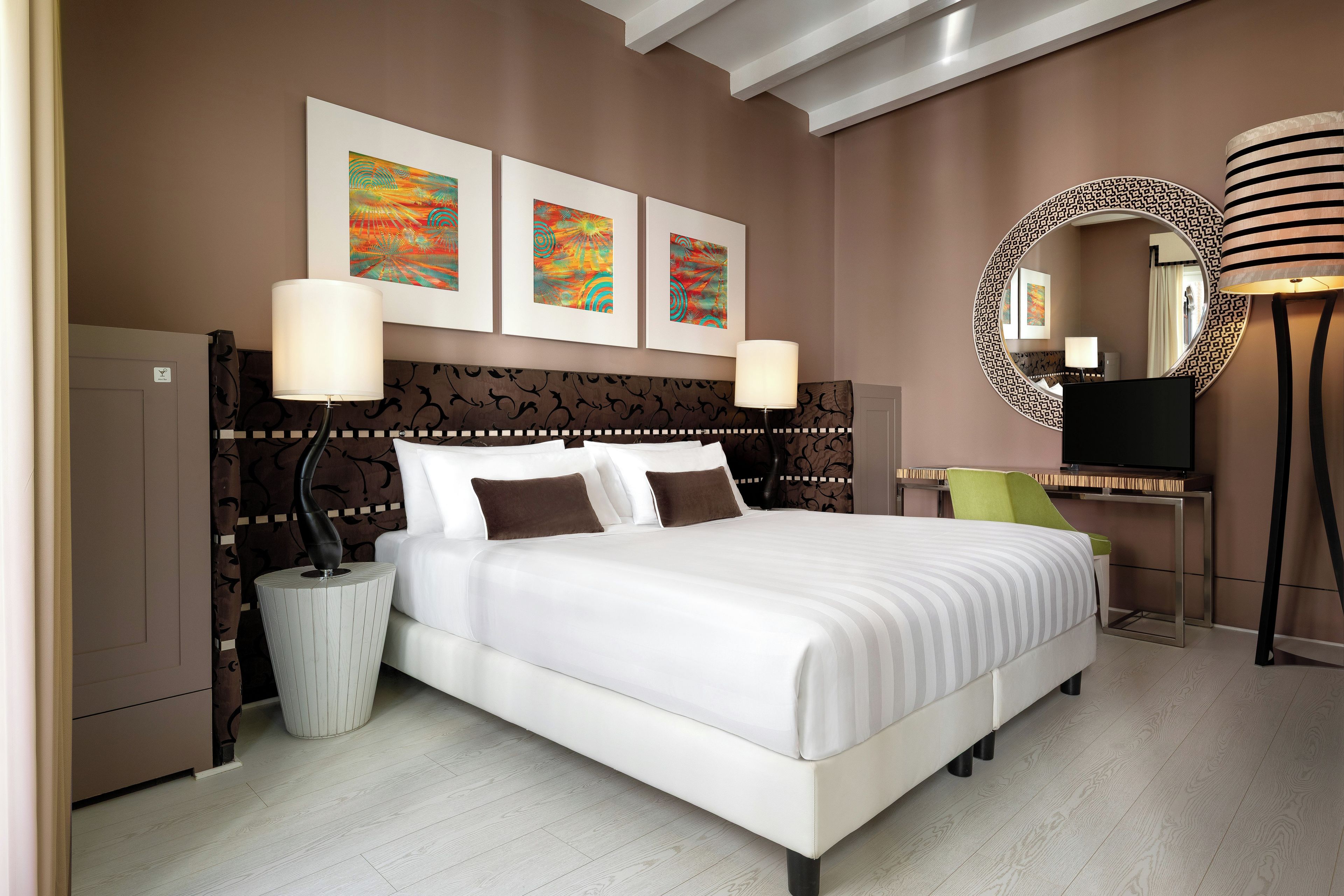 A hotel room with a large bed, two bedside lamps, a round mirror, a TV, and three framed artworks on the wall.
