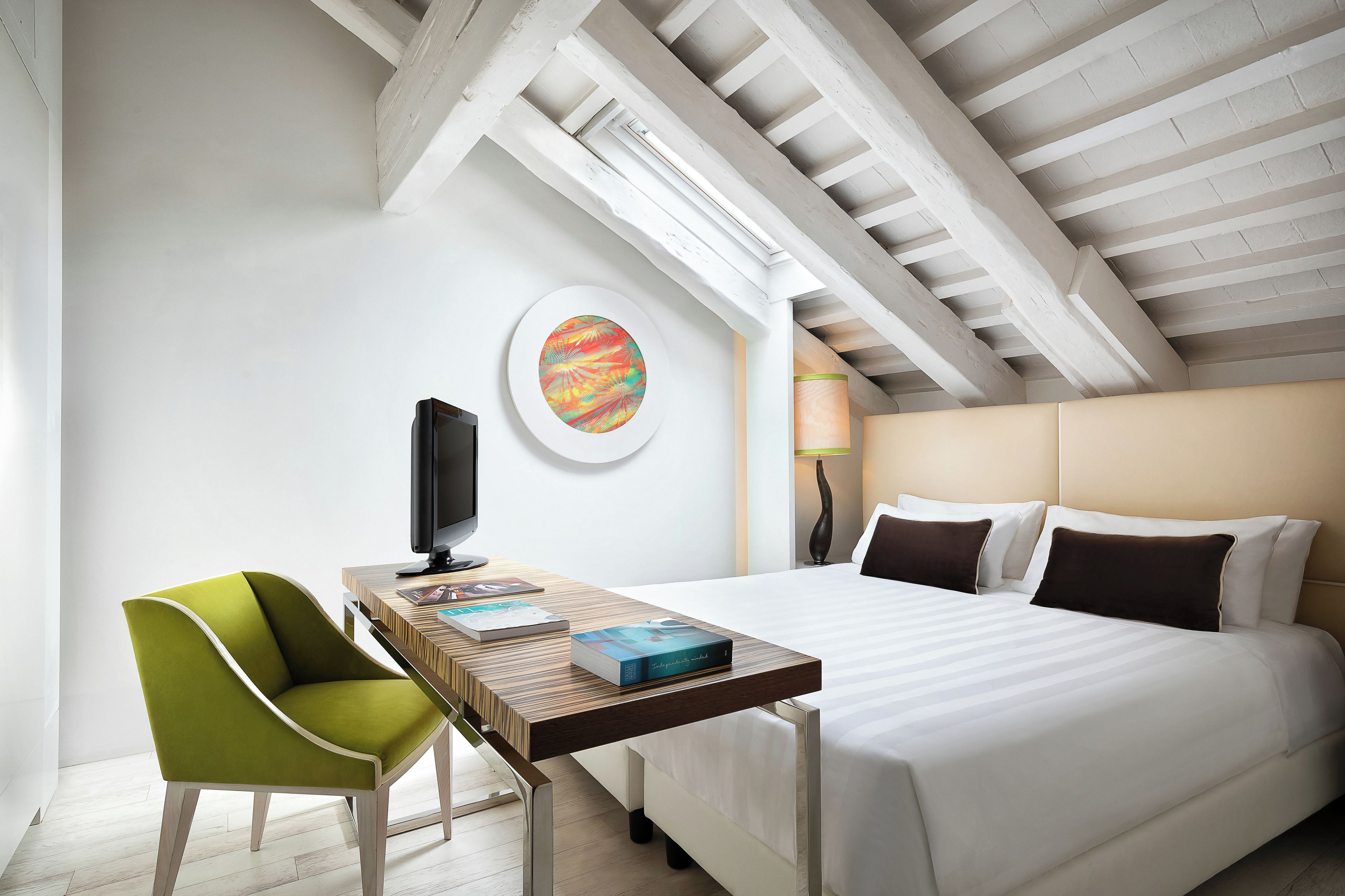 A hotel room with a bed, a desk with a computer, a chair, a lamp, and a wall art piece.