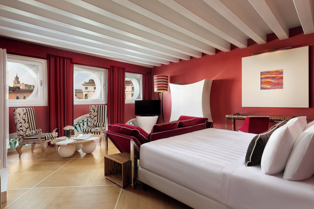 Habitación de hotel con una cama grande, dos sillones, una mesita y vistas de la ciudad a través de amplios ventanales.