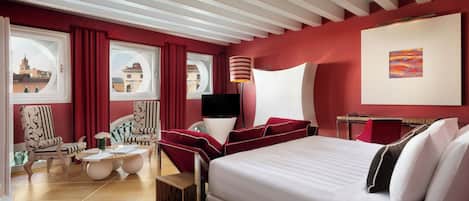 Habitación de hotel con una cama grande, dos sillones, una mesita y vistas de la ciudad a través de amplios ventanales.