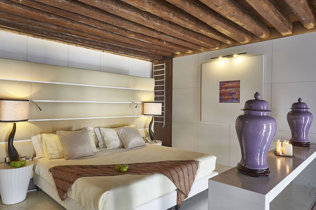 Un dormitorio moderno con techo de madera, una cama con ropa blanca, una mesita de noche con lámpara y jarrones decorativos sobre una consola.