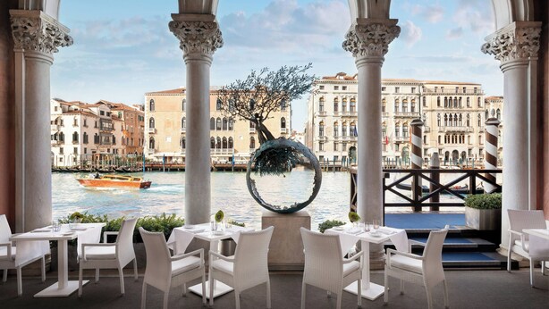 Un restaurante con sillas y mesas de mimbre blanco, vista a un canal con edificios, y un objeto esférico decorativo.