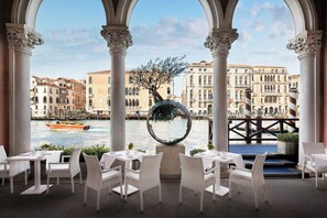 A restaurant with white wicker chairs and tables, a view of a canal with buildings, and a decorative spherical object. - Sina Centurion Palace (Venice)