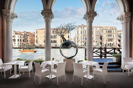 A restaurant with white wicker chairs and tables, a view of a canal with buildings, and a decorative spherical object.