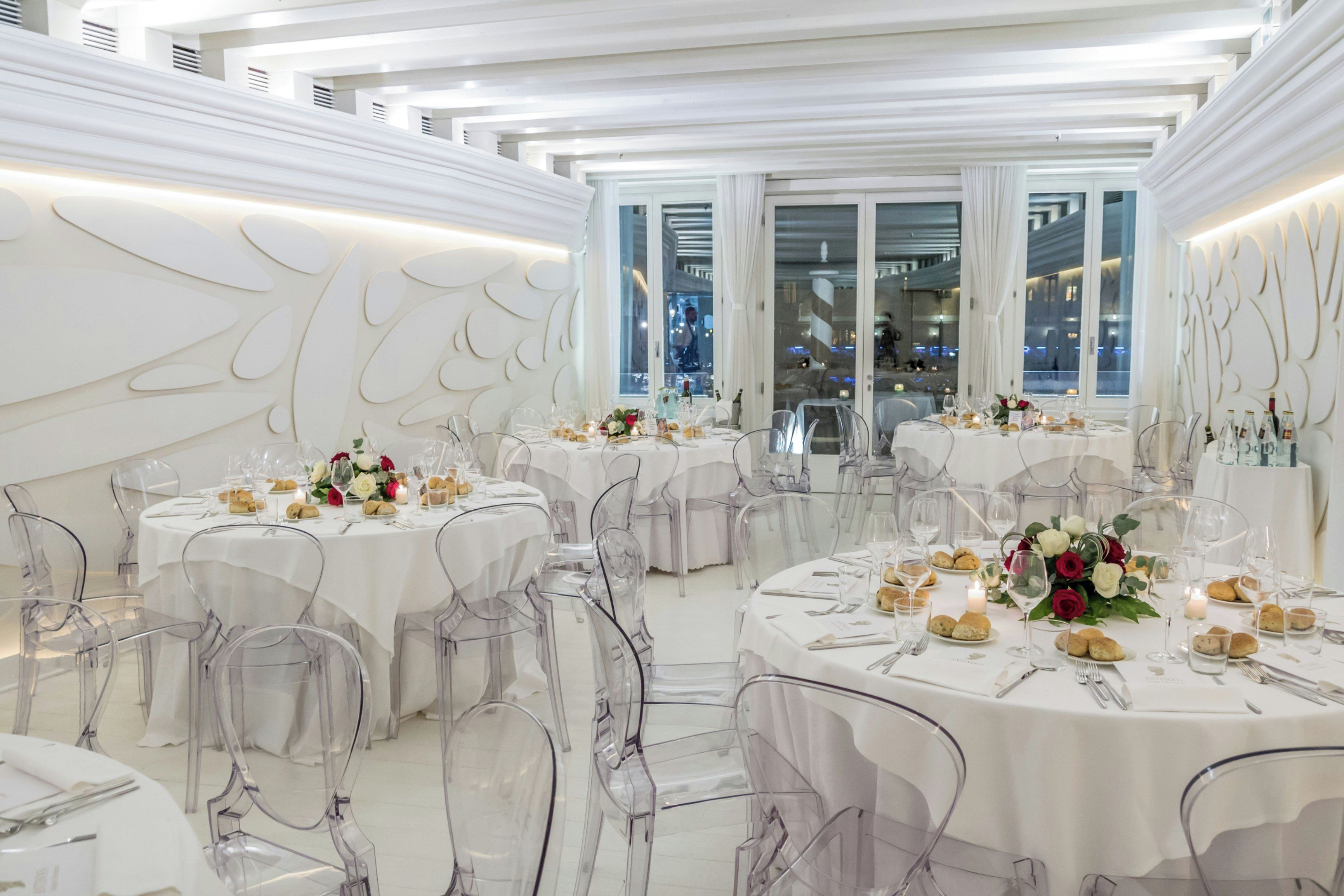 A spacious, elegantly decorated dining area with round tables, clear chairs, and white drapery.