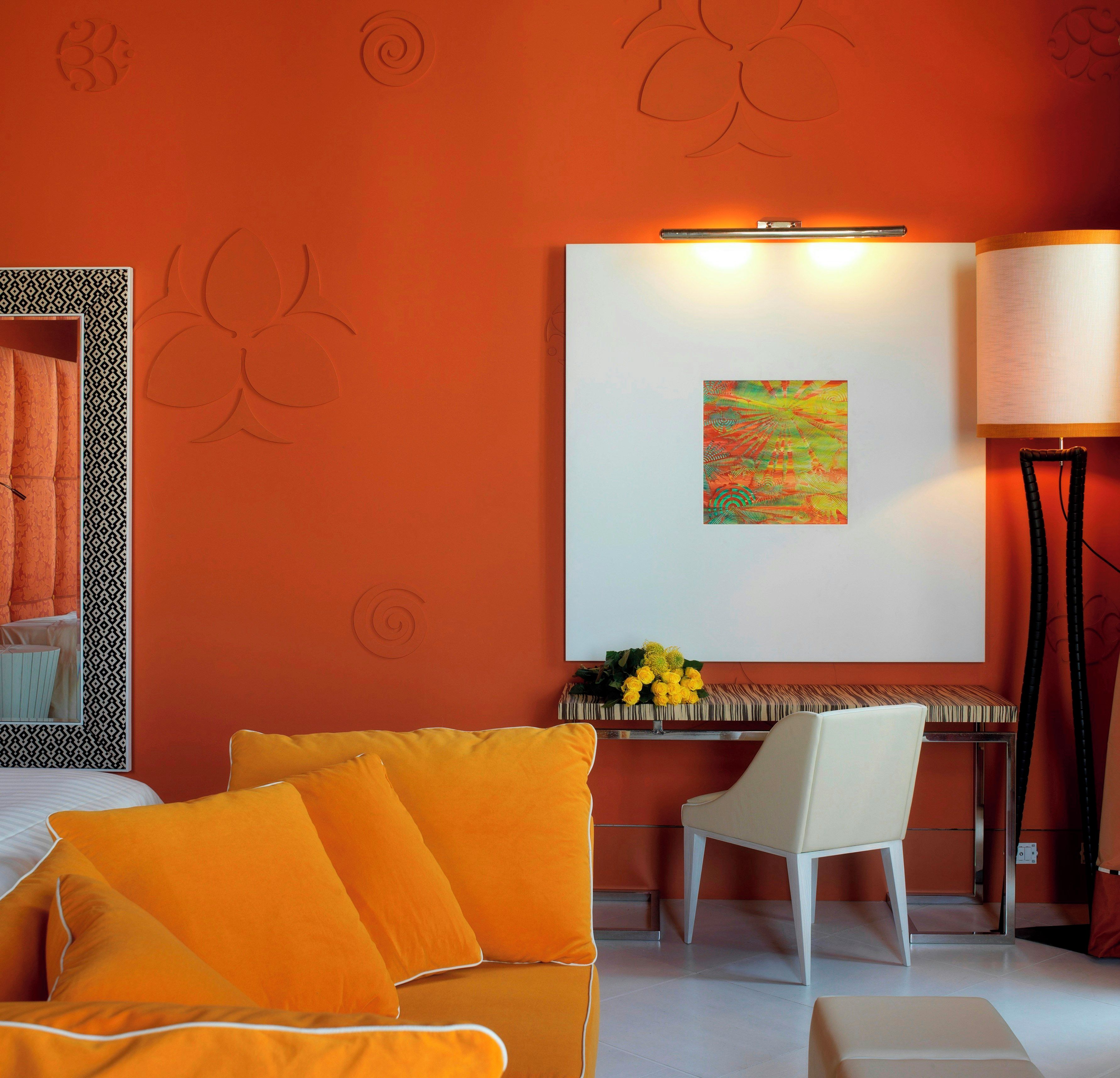 Una habitación de hotel con una pared naranja, una cama con ropa blanca, un sofá amarillo, una silla blanca, un escritorio con una lámpara y un cuadro en la pared.