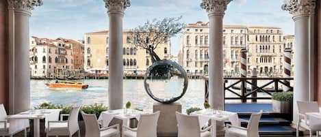 A restaurant with white wicker chairs and tables, a view of a canal with buildings, and a decorative spherical object.