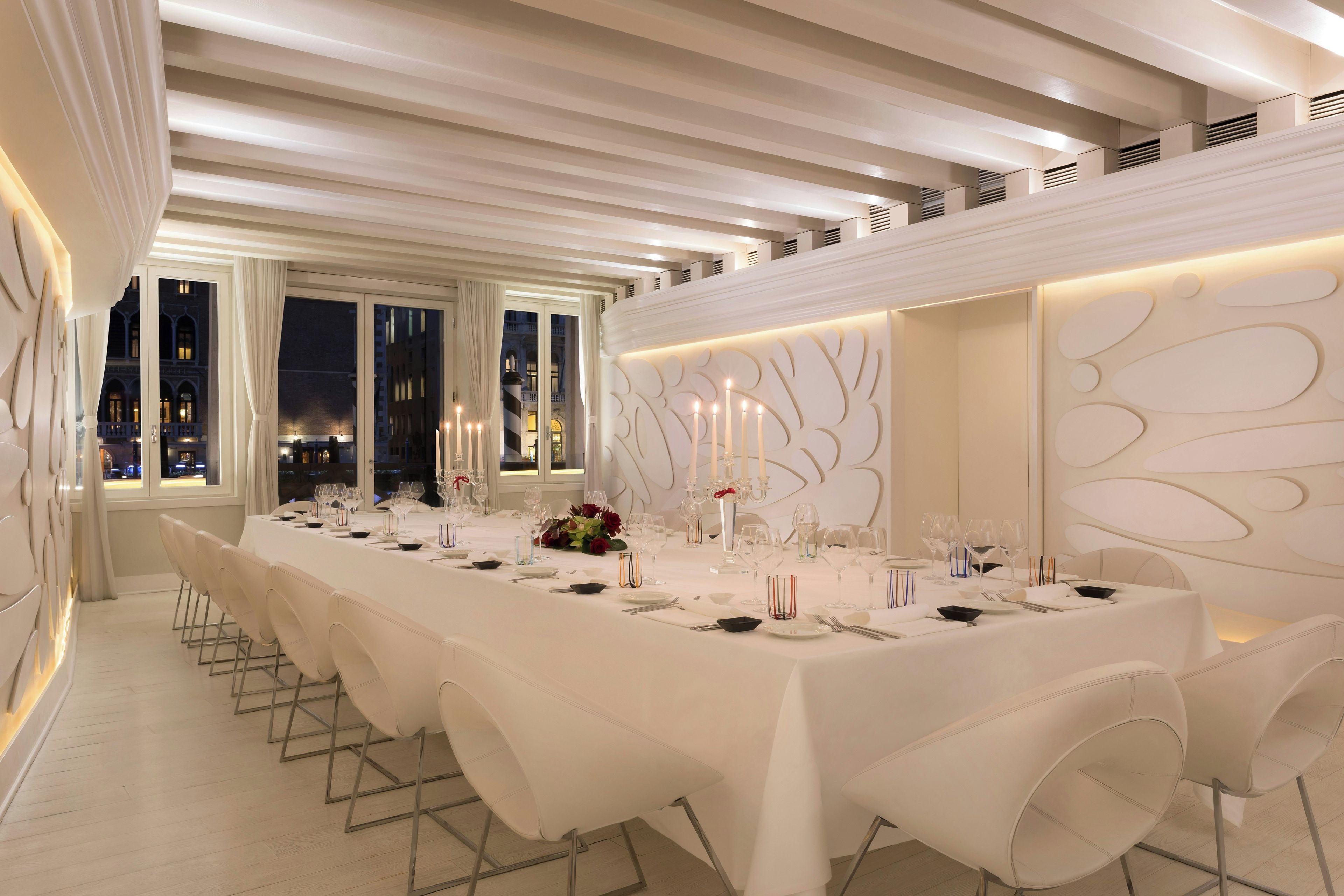 A long dining table set for a formal meal, with white tablecloths, candles, and modern chairs.