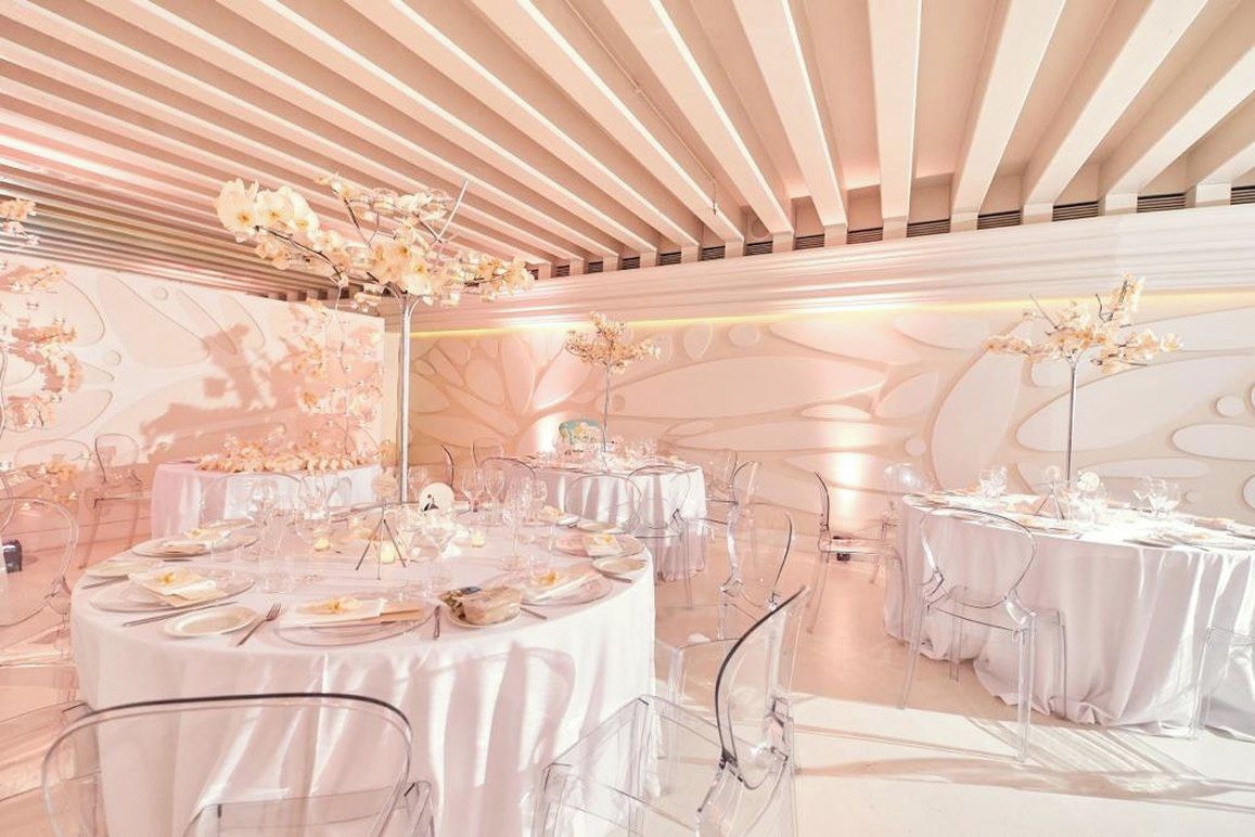 Un espacio para eventos amplio y elegantemente decorado, con mesas redondas, sillas transparentes y arreglos florales.