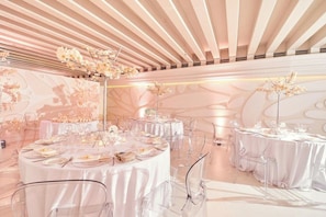 Um espaço de eventos amplo e elegantemente decorado, com mesas redondas, cadeiras transparentes e arranjos florais.