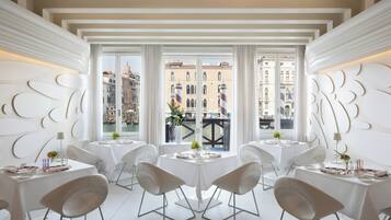 Un espace de restauration moderne avec des tables et des chaises blanches, de grandes fenêtres offrant une vue sur la ville et des panneaux décoratifs sur les murs.