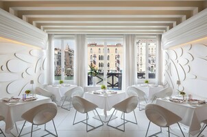 Un comedor moderno con mesas y sillas blancas, ventanas amplias con vista a la ciudad y paneles decorativos en la pared.