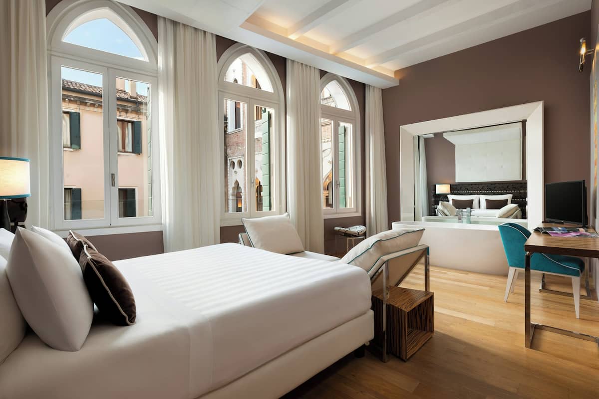 Habitación de hotel con cama, escritorio, silla y ventanas amplias.