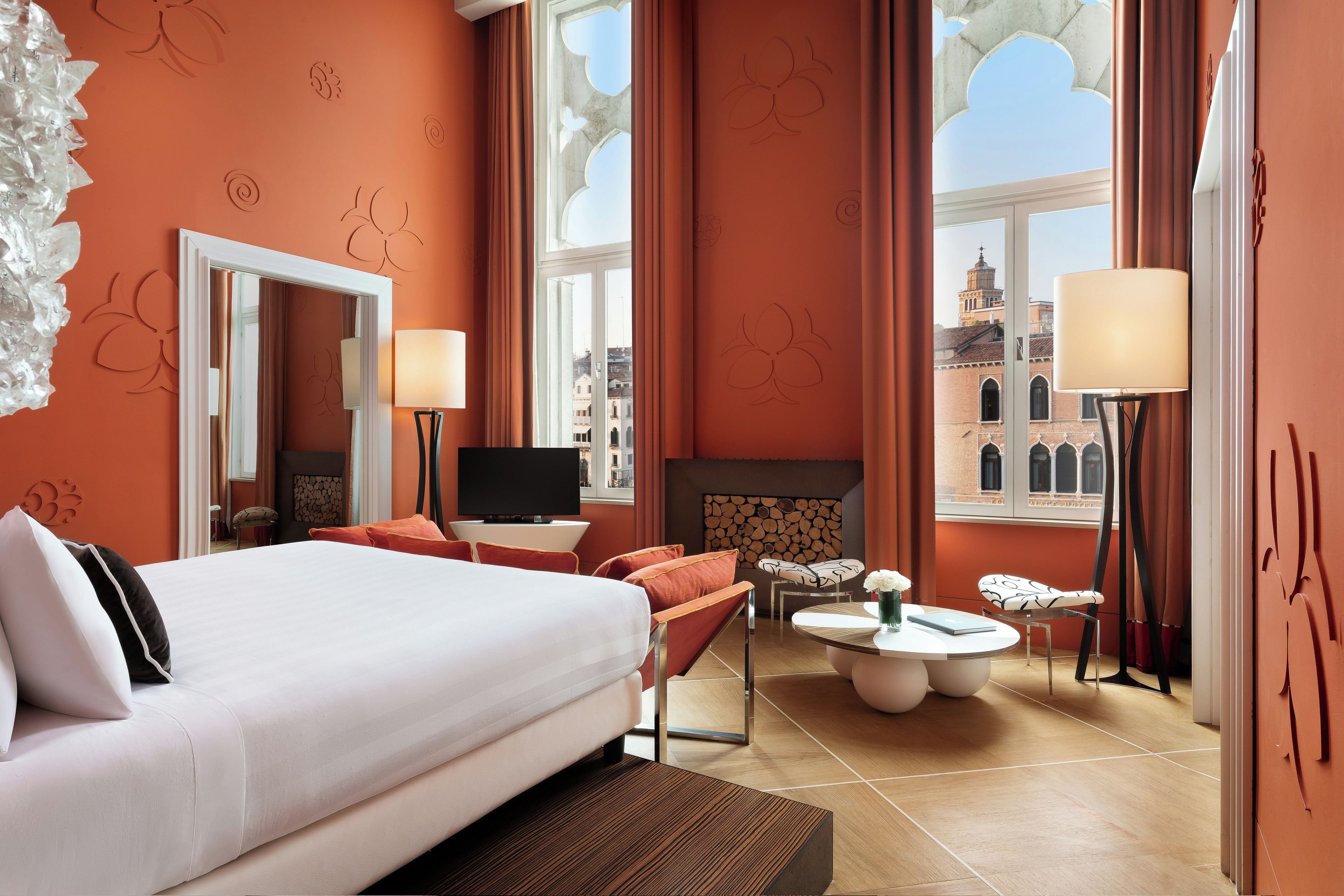 Habitación de hotel con una cama grande, televisor, zona de estar con mesa y vistas a la ciudad a través de amplios ventanales.