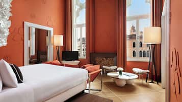 Habitación de hotel con una cama grande, televisor, zona de estar con mesa y vistas a la ciudad a través de amplios ventanales.