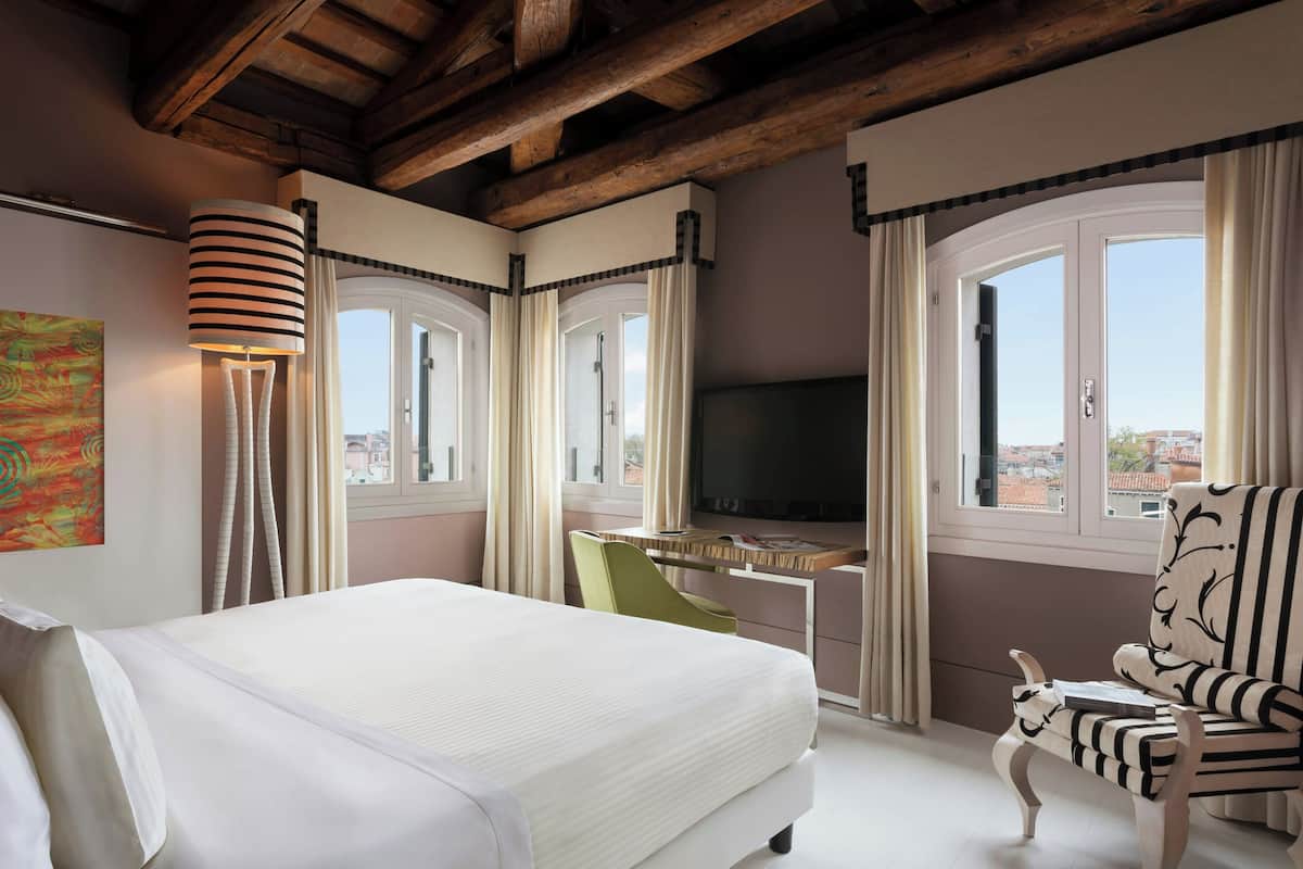 Una habitación de hotel con cama, escritorio con televisor, silla y ventanas amplias.