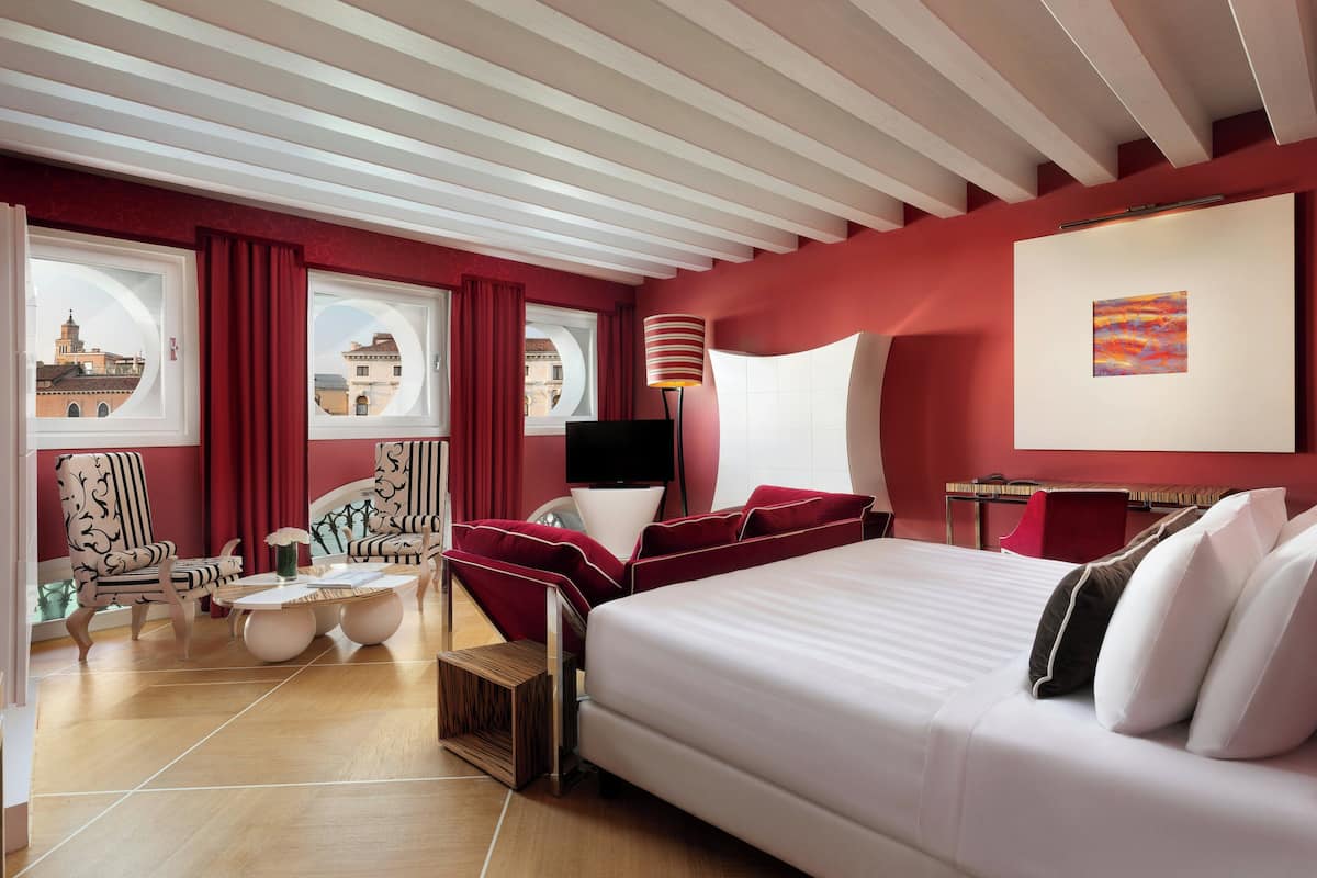 Habitación de hotel con una cama grande, dos sillones, una mesita y vistas de la ciudad a través de amplios ventanales.