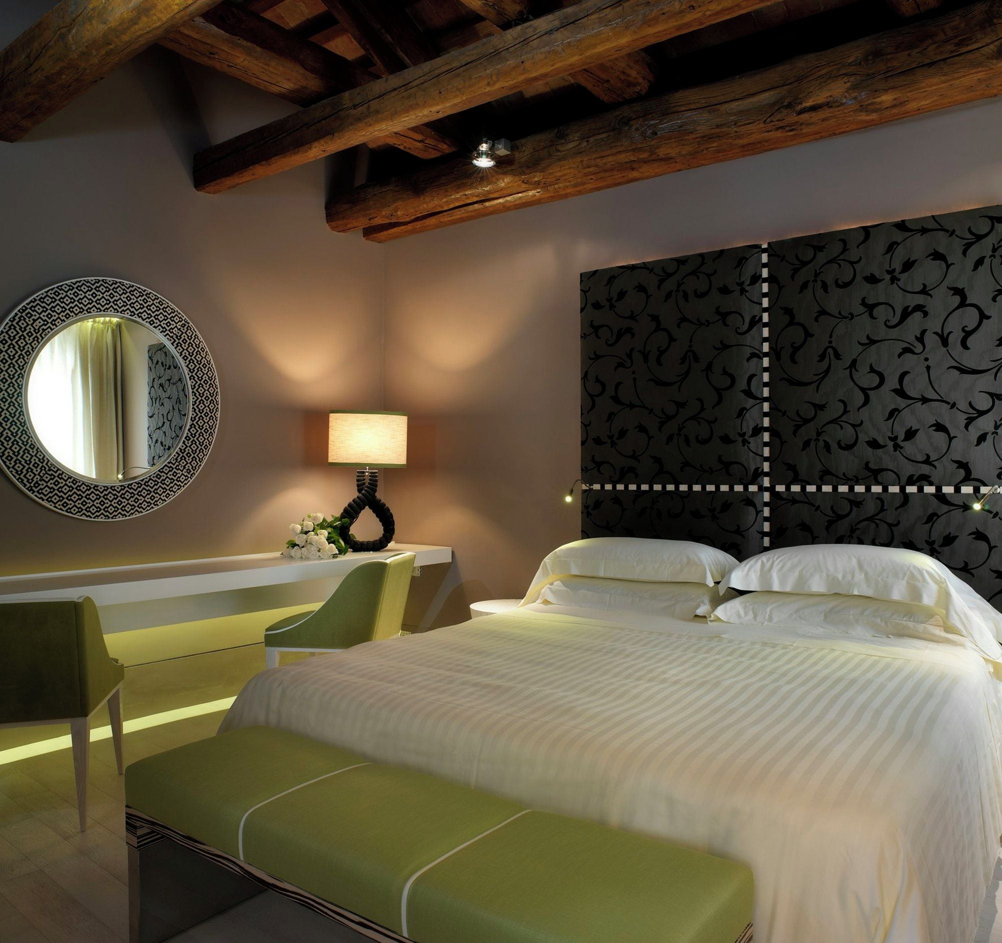 A hotel room with a large bed, a round mirror, a bedside table with a lamp, and a green bench.