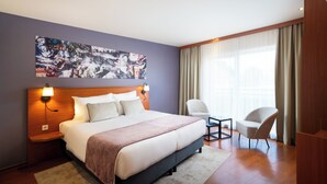 A modern hotel room with a large bed, a nightstand with a lamp, a chair, and a large window with curtains. -  Leonardo Hotel Salzburg City Center (Salzburg)