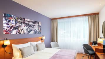 Ein Hotelzimmer mit einem großen Bett, einem Schreibtisch, einem Stuhl, einer Schreibtischlampe, einem an der Wand montierten Fernseher und einem Fenster mit Vorhängen.