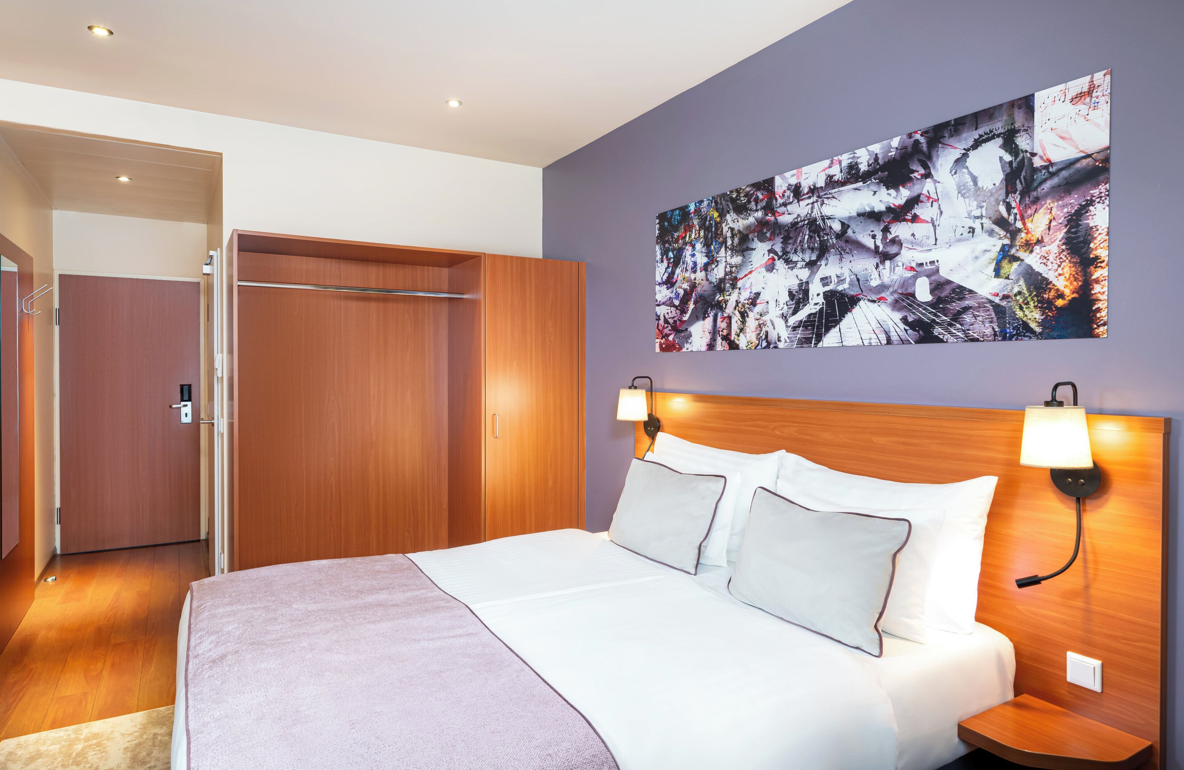 A modern hotel room with a large bed, wooden wardrobe, and a wall-mounted artwork.