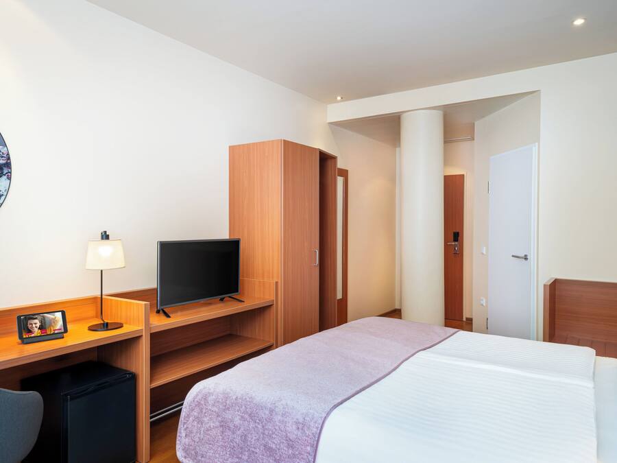 Quarto de hotel com cama, escrivaninha com televisão, luminária e pequeno frigorífico.