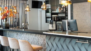 A modern bar area with a bar counter, three bar stools, hanging pendant lights, and a television screen above the counter. -  Leonardo Hotel Salzburg City Center (Salzburg)