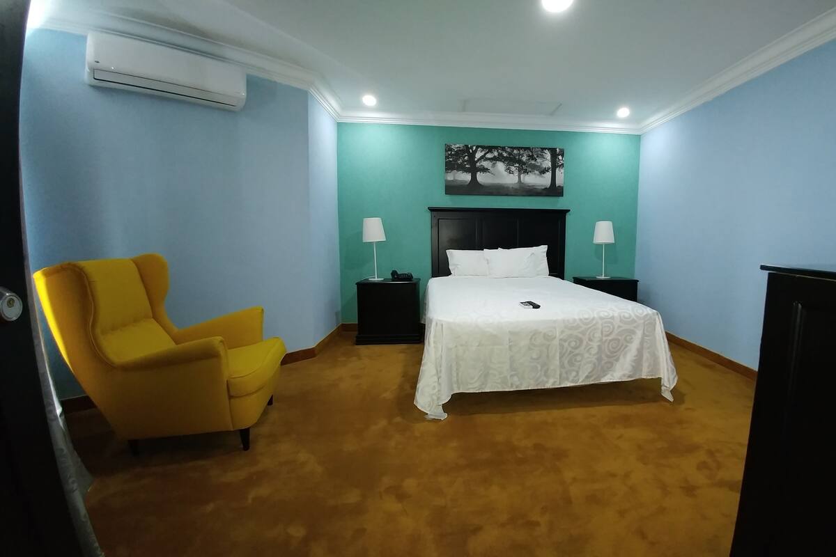 Een hotelkamer met een bed, een gele fauteuil, twee nachtkastjes met lampen, een schilderij aan de muur en een airconditioning.
