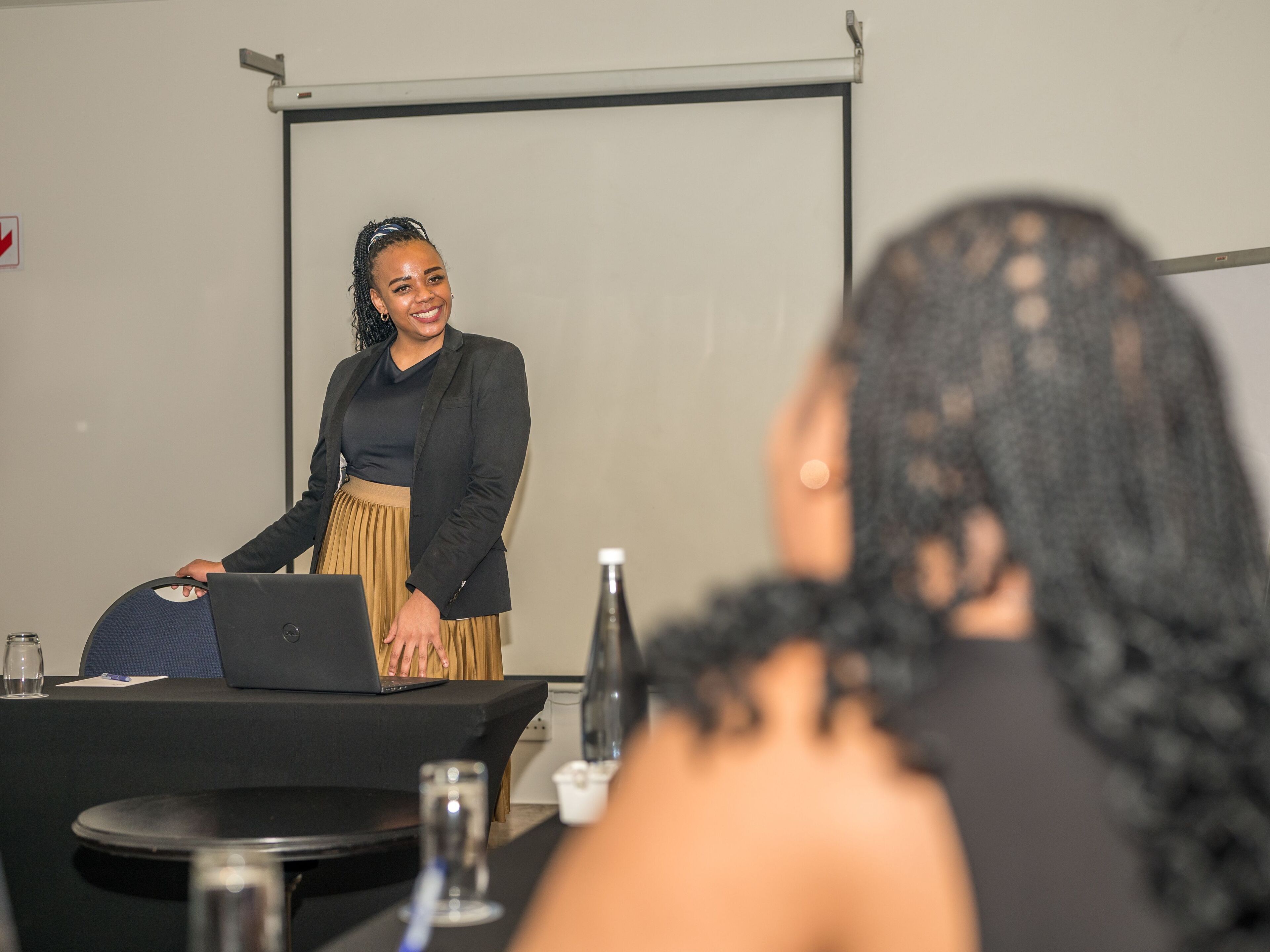 A person presenting at a table with a laptop, a projector screen, and a water bottle in the background.