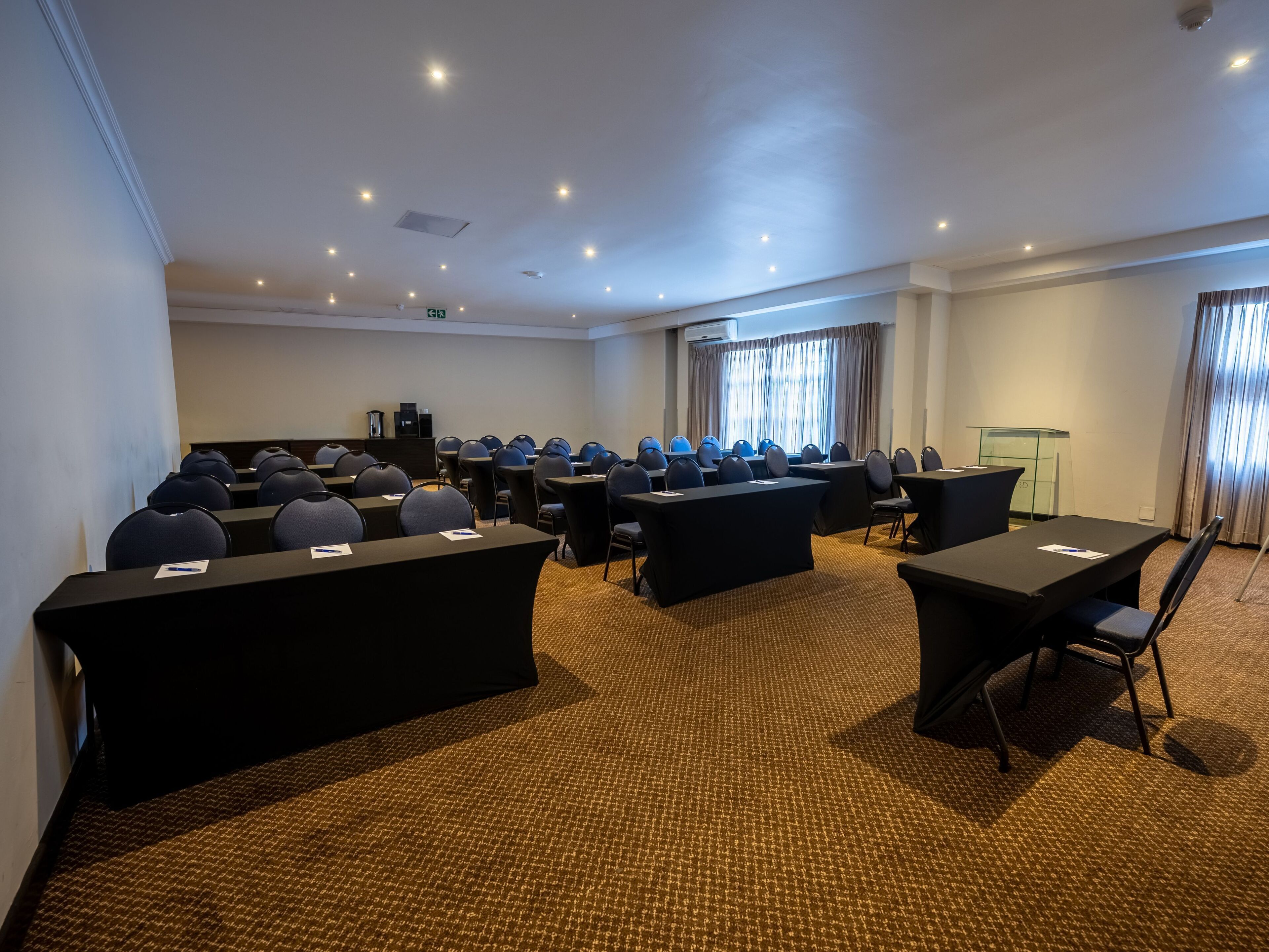Un salón de conferencias con filas de mesas y sillas, un atril y un ventanal con cortinas.
