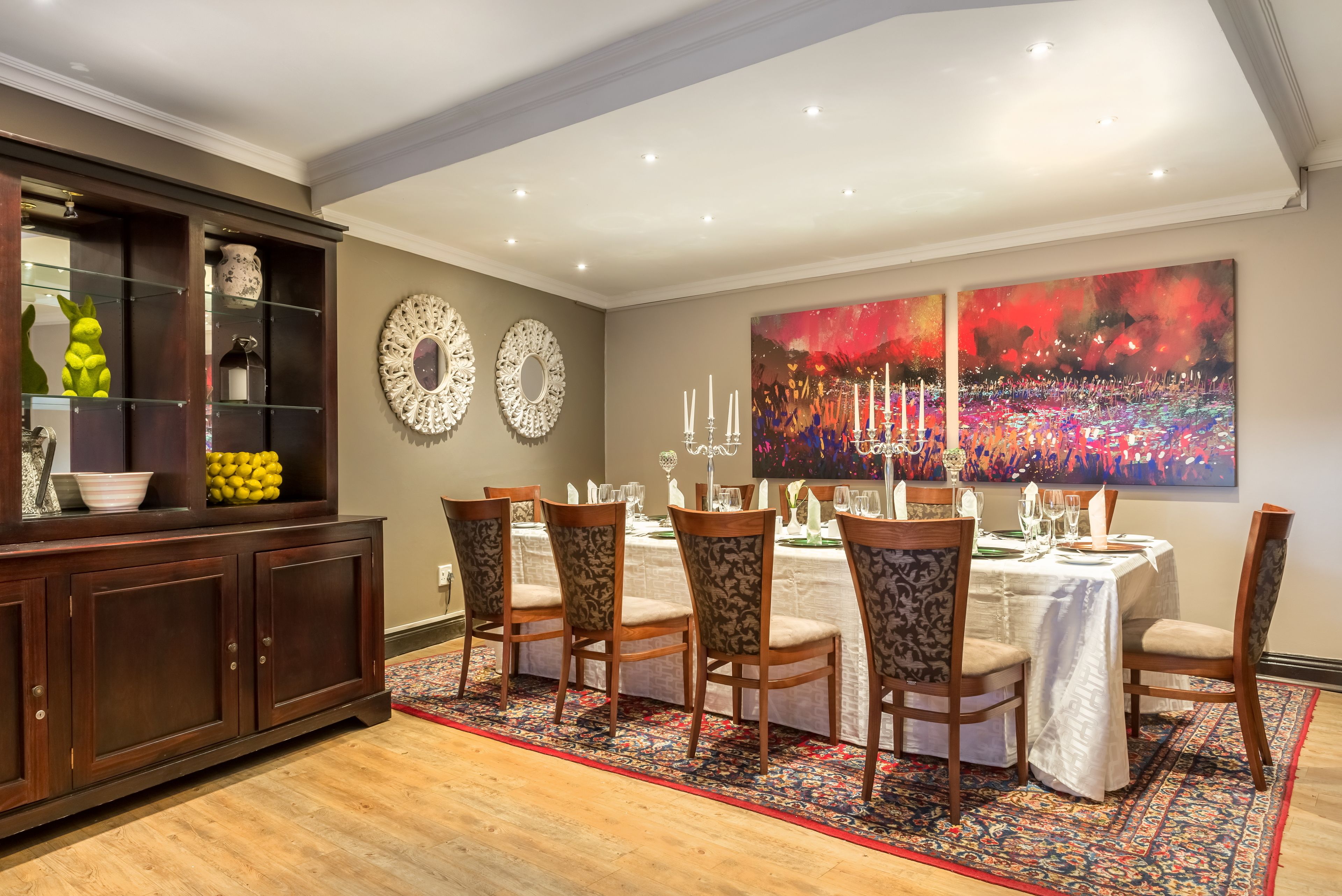 A dining area with a table set for six, surrounded by patterned chairs, a dark wooden cabinet with glass doors, and two decorative wall mirrors.