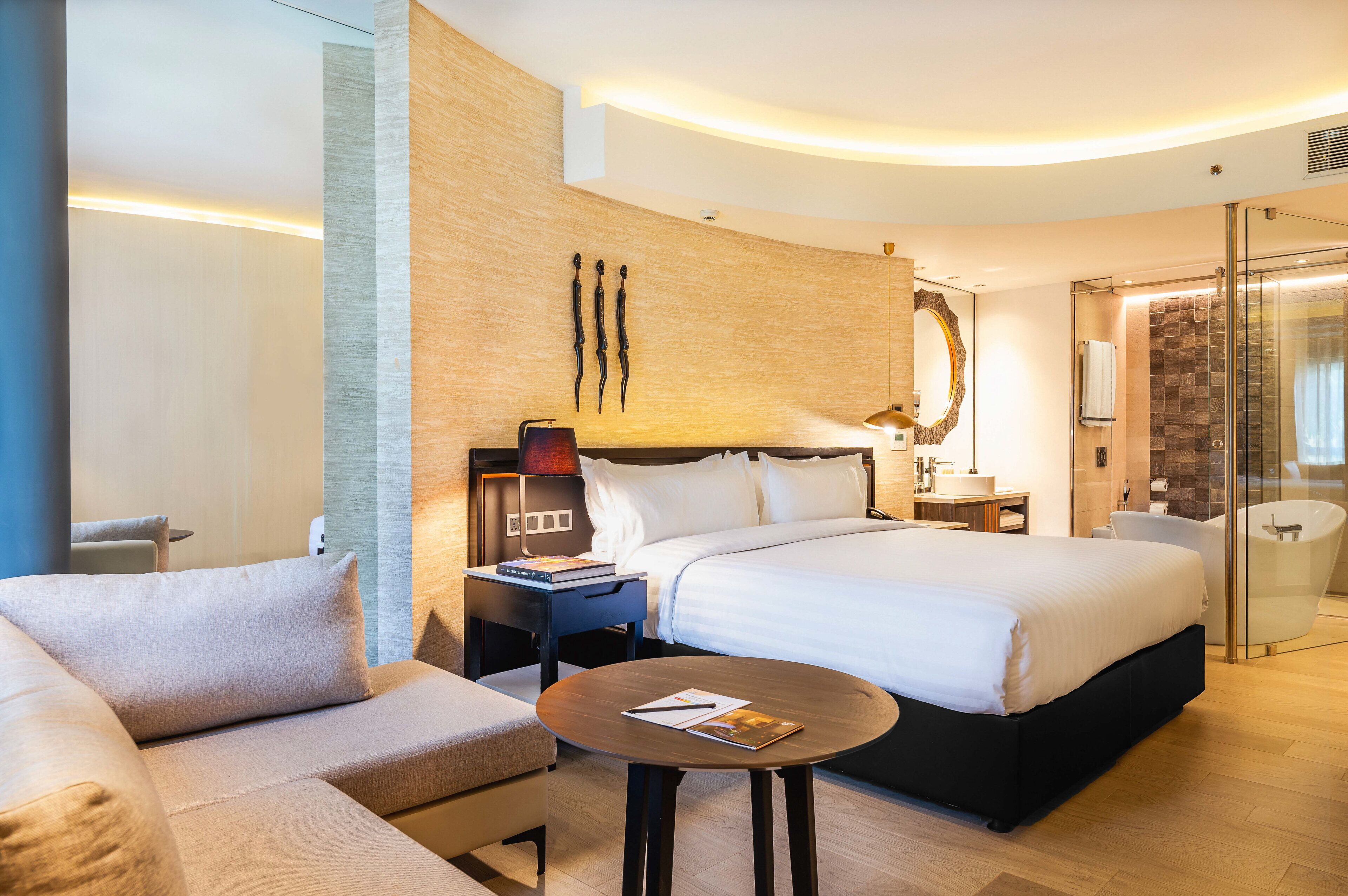 A modern hotel room with a large bed, a sofa, a bedside table with a lamp, a glass shower enclosure, and a wooden accent wall.
