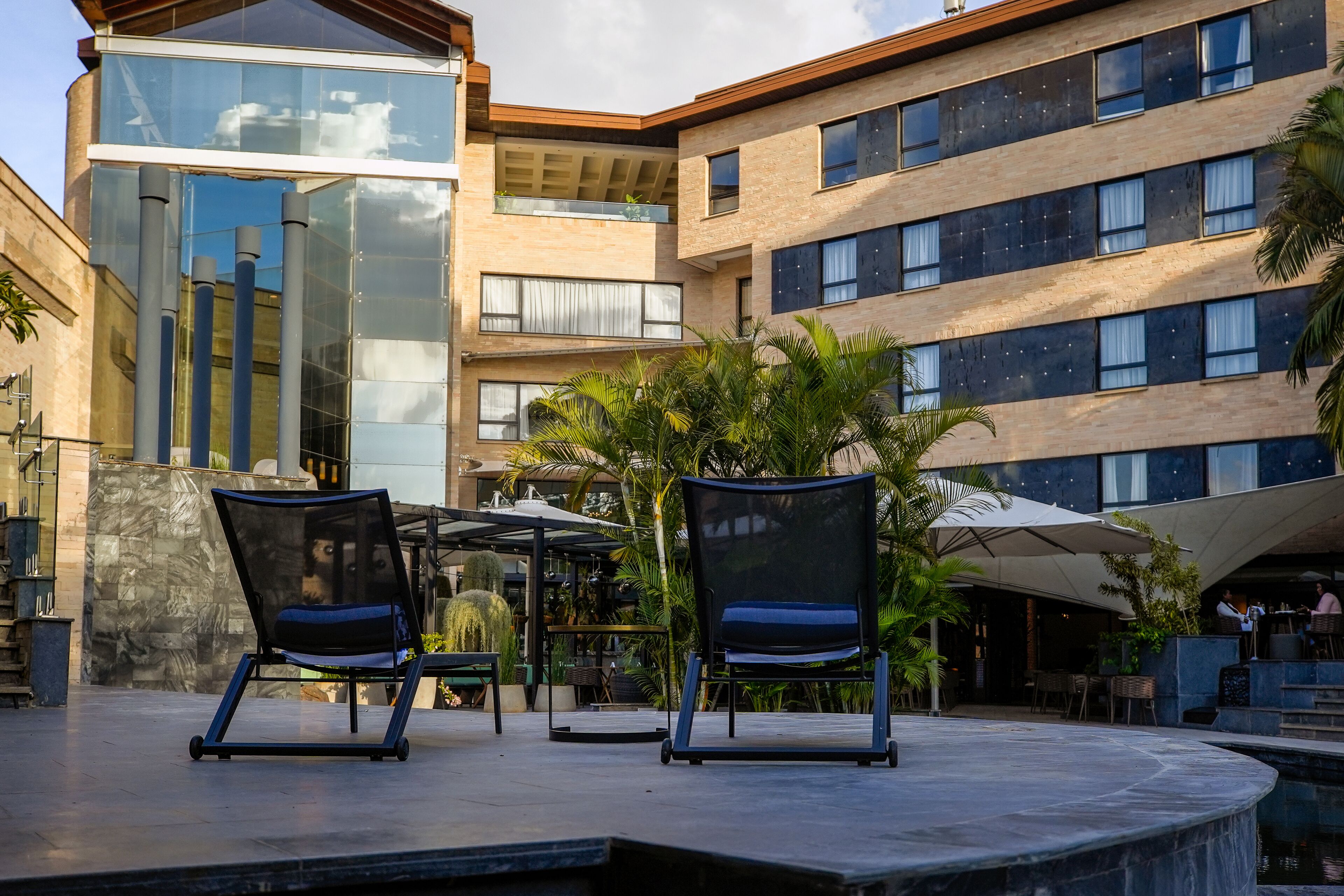 Un edificio moderno de varios pisos, con amplios ventanales y un patio con mobiliario exterior y palmeras.