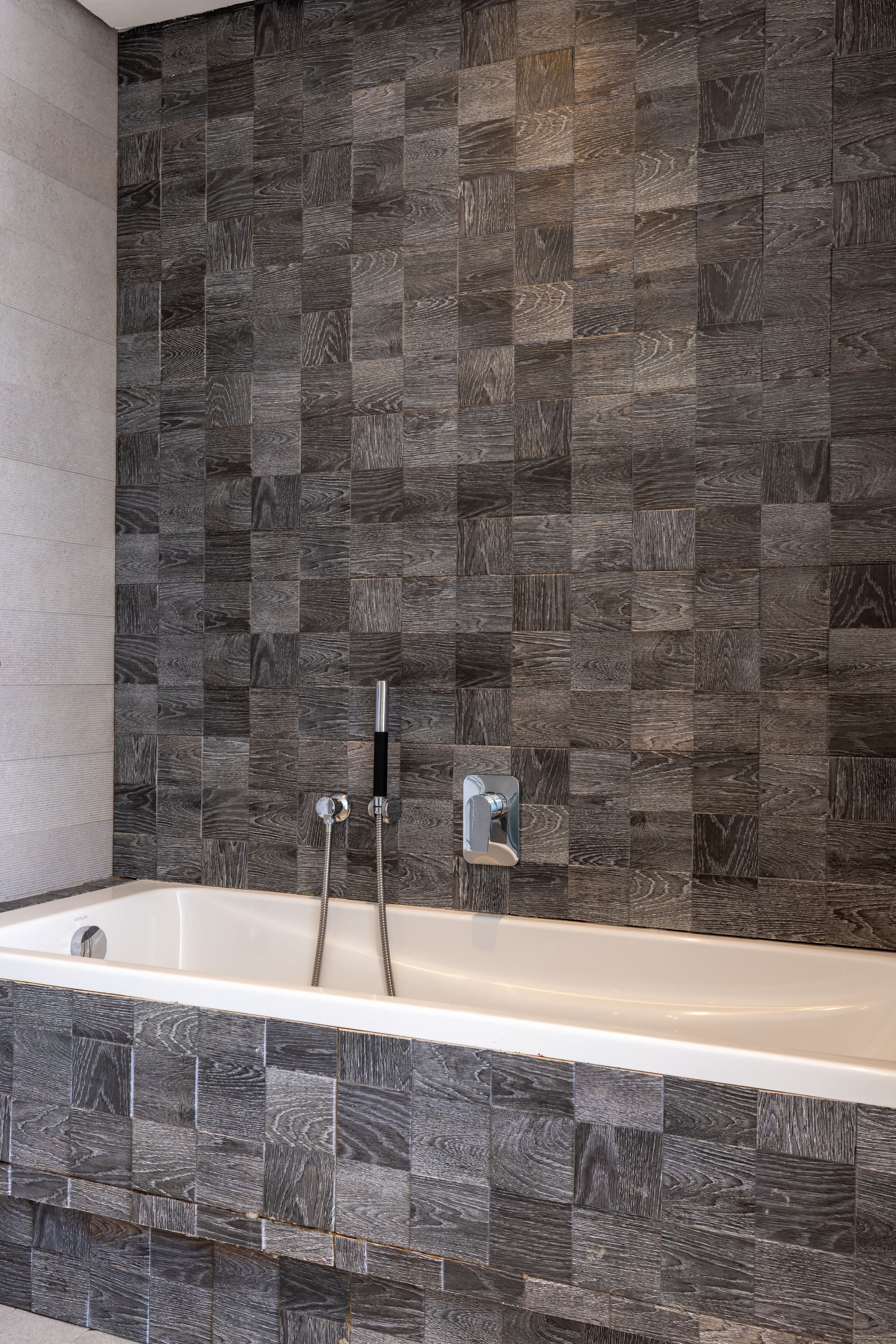A modern bathroom with a large bathtub, dark wood-patterned tiles, and a chrome showerhead.