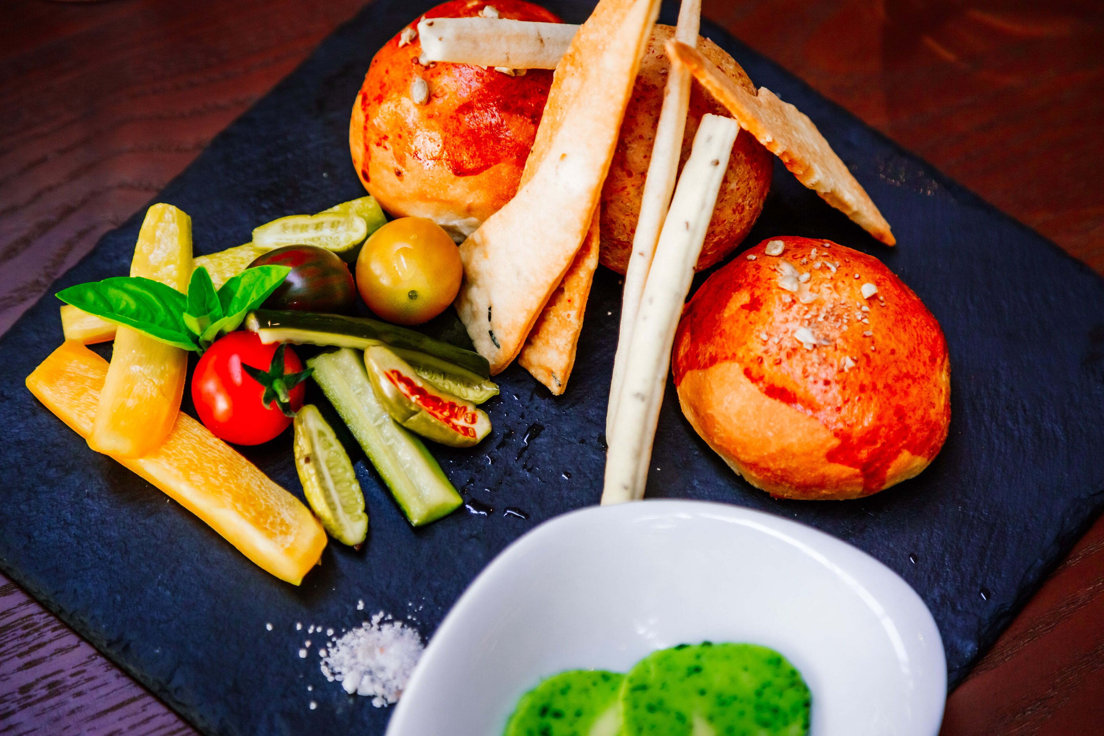 A dark slate board with a variety of food items including bread, olives, tomatoes, and pickles.