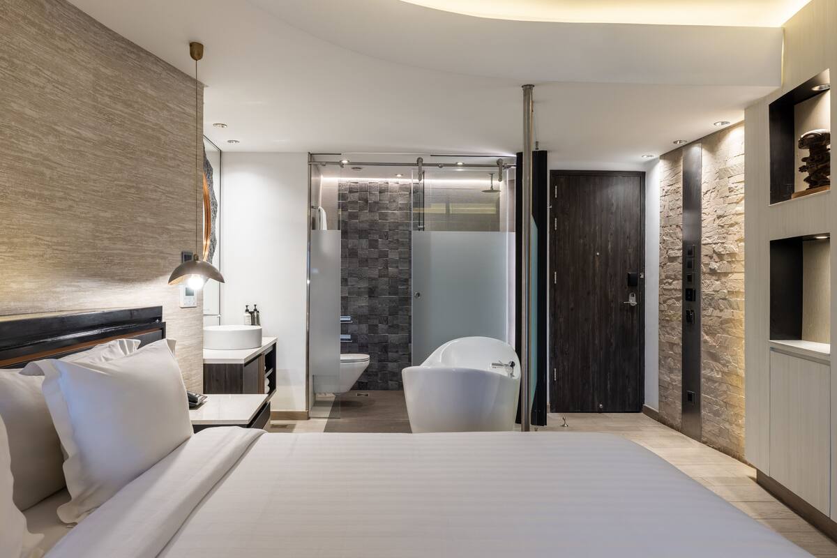 Moderni hotellihuone, jossa on suuri sänky, kylpyhuone lasiseinällä varustetulla suihkulla sekä yksi seinä, joka on koristeltu kivellä.