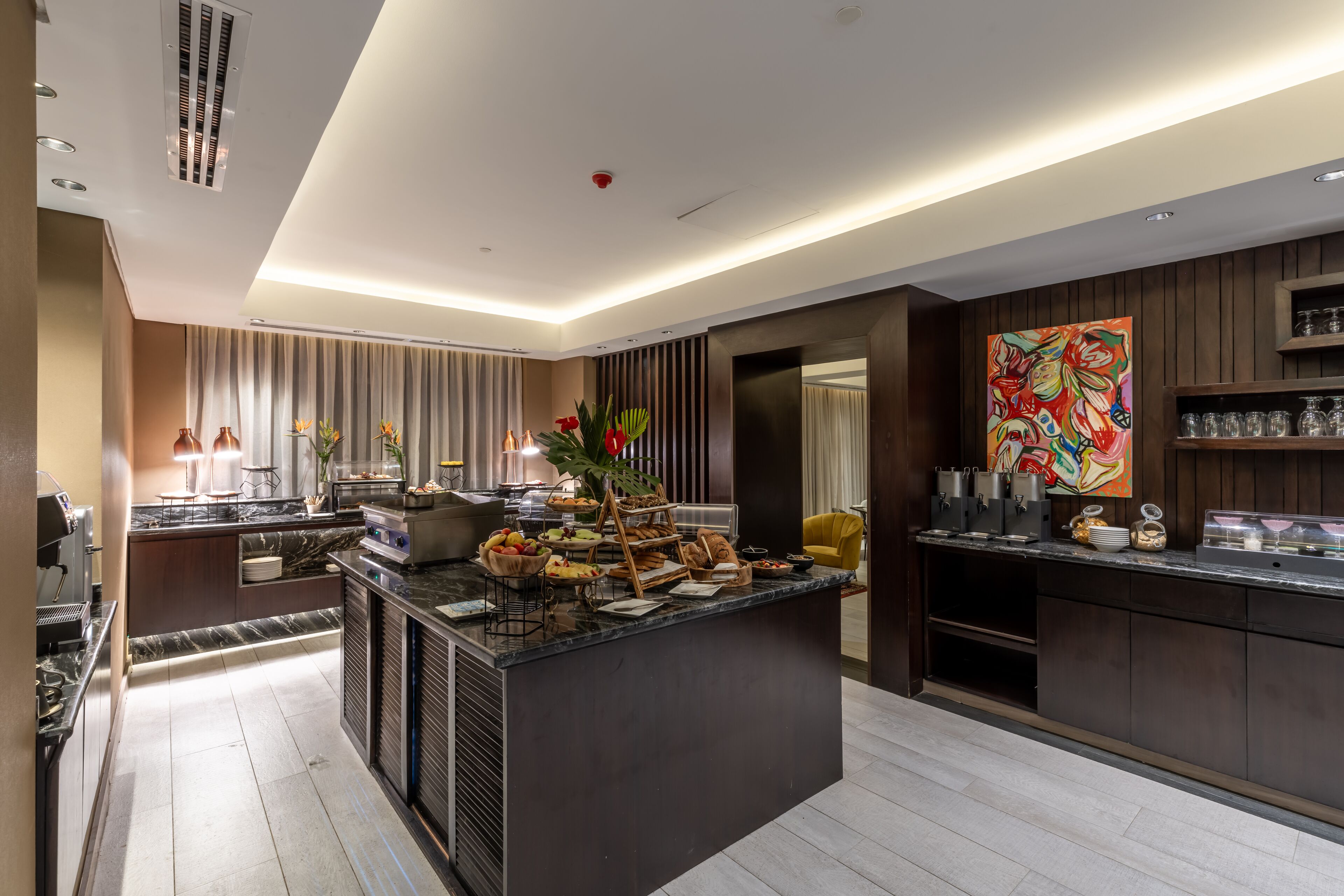 A modern kitchen with a marble countertop, a central island with food and drinks, and a large abstract painting on the wall.