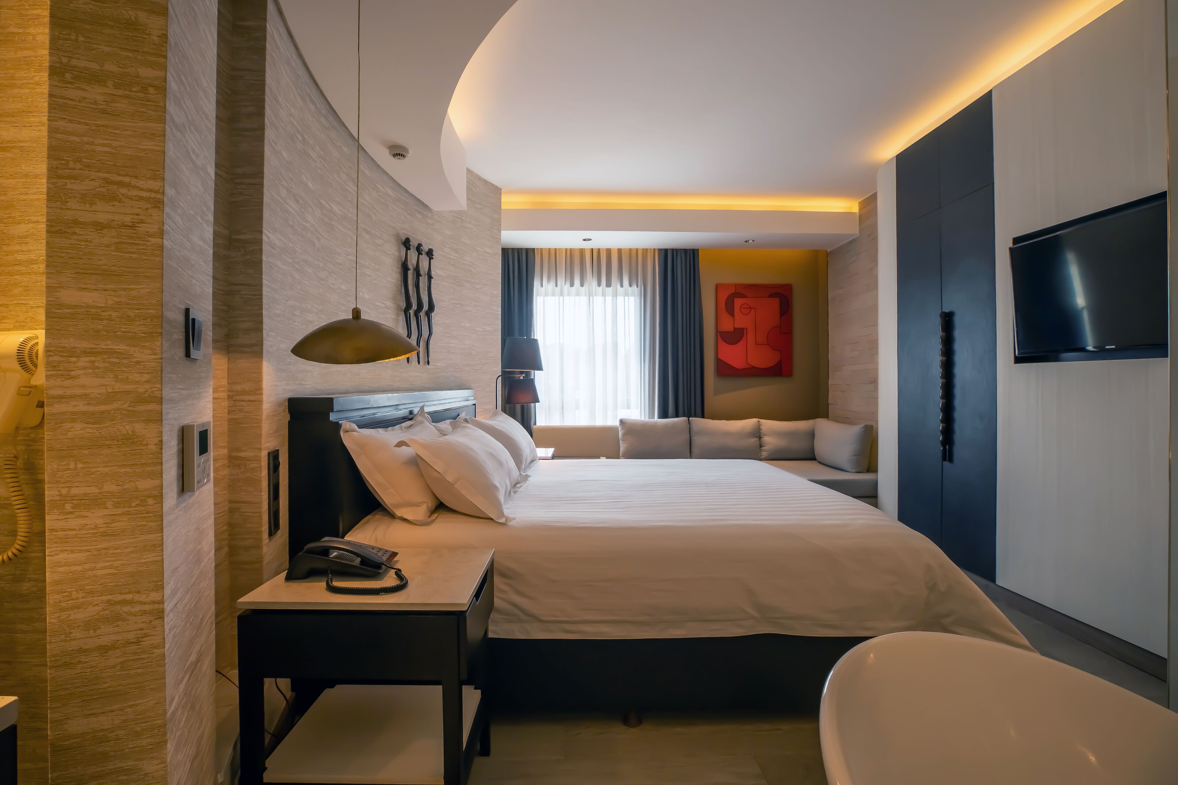 A modern hotel room with a large bed, a desk with a telephone, a bedside lamp, a TV mounted on the wall, and a bathtub.
